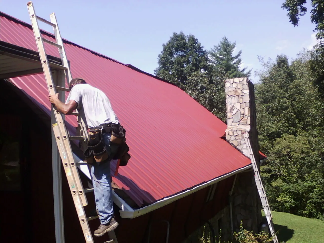 Roofer on a ladder beside a red metal roof and stone chimney — Soffit & Fascia Repair by Roofing Brothers in Rawlings